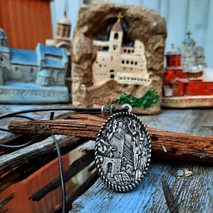 May include: A silver pendant necklace featuring a detailed image of a church and figures. The pendant is oval shaped and hangs from a black leather cord. The pendant is displayed in front of a wooden surface and several miniature church models.