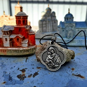 May include: A silver pendant necklace with a depiction of the Virgin Mary holding baby Jesus. The pendant is on a black cord and is resting on a small rock. The background features miniature models of churches.