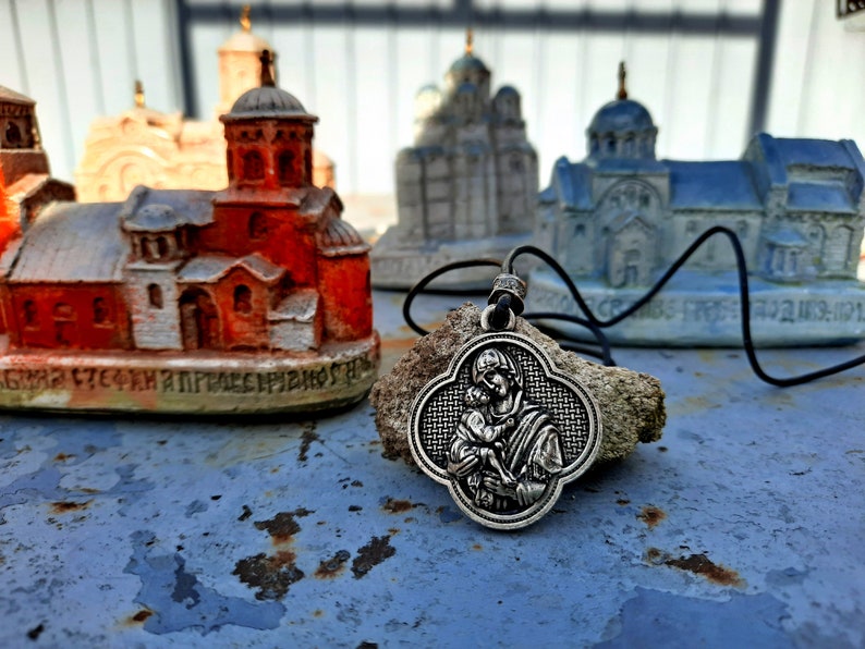 May include: A silver pendant necklace with a depiction of the Virgin Mary holding baby Jesus. The pendant is on a black cord and is resting on a small rock. The background features miniature models of churches.