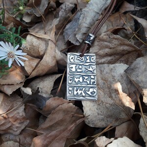 May include: A silver pendant necklace with a brown leather cord. The pendant features a rectangular design with an inscription of symbols and characters. The necklace is lying on a bed of brown leaves.