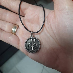 May include: A silver pendant necklace with a black cord. The pendant features a circular design with the Greek letters 'alpha' and 'omega' and the letter 'P' in the center.