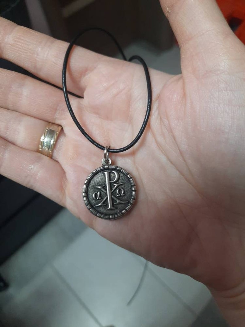 May include: A silver pendant necklace with a black cord. The pendant features a circular design with the Greek letters 'alpha' and 'omega' and the letter 'P' in the center.
