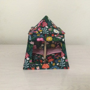 May include: A small, fabric play tent with a dark green background and a colorful floral and mushroom pattern. The tent has a pink interior and is supported by wooden dowels. The tent is designed for children's play.