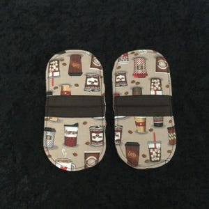 Pode incluir: Duas luvas de forno com um padrão de tema de café. As luvas bege apresentam um design repetido de xícaras de café, grãos de café e as palavras "Espresso" e "Latte". Cada luva tem uma faixa marrom escuro no meio.