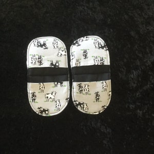 May include: A pair of white oven mitts with a black cow print design. The mitts have a black band around the middle.