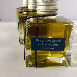 Pode incluir: Quatro pequenas garrafas de vidro de azeite extra virgem grego premium com tampas prateadas e um cord&atilde;o azul amarrado ao redor do pesco&ccedil;o. As garrafas est&atilde;o sobre uma superf&iacute;cie branca.