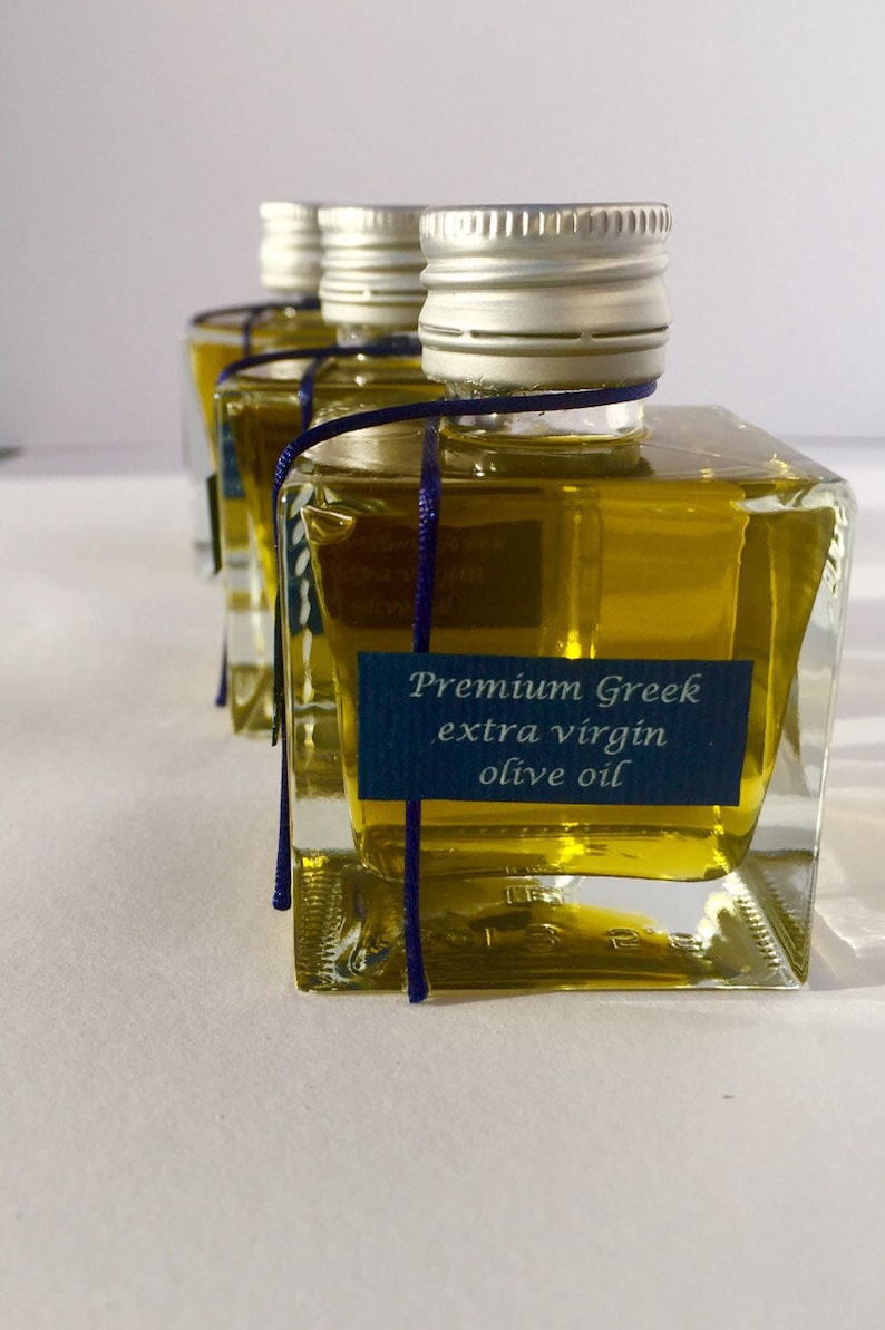 Pode incluir: Quatro pequenas garrafas de vidro de azeite extra virgem grego premium com tampas prateadas e um cord&atilde;o azul amarrado ao redor do pesco&ccedil;o. As garrafas est&atilde;o sobre uma superf&iacute;cie branca.