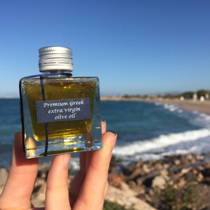 Pode incluir: Uma pequena garrafa de vidro de azeite extra virgem grego premium, segurada na m&atilde;o contra um fundo de mar azul e praia de areia.
