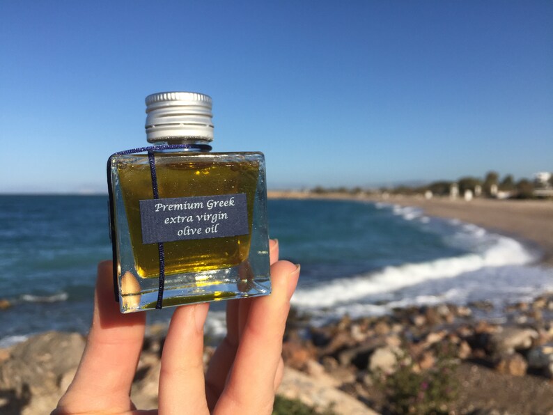 Pode incluir: Uma pequena garrafa de vidro de azeite extra virgem grego premium, segurada na m&atilde;o contra um fundo de mar azul e praia de areia.