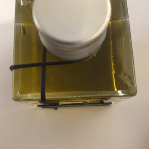 Pode incluir: Uma garrafa de vidro transparente com uma tampa prateada e um cord&atilde;o preto amarrado ao redor do pesco&ccedil;o. A garrafa est&aacute; cheia de um l&iacute;quido dourado.