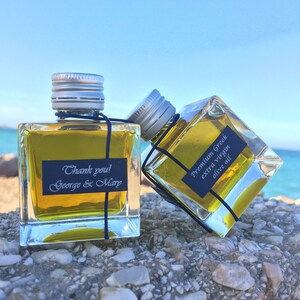 Pode incluir: Duas pequenas garrafas de vidro de azeite de oliva com tampas prateadas. As garrafas est&atilde;o sentadas em uma superf&iacute;cie rochosa. Uma garrafa tem um r&oacute;tulo que diz "Obrigado! George & Mary". A outra garrafa tem um r&oacute;tulo que diz "Azeite de oliva extra virgem grego premium".