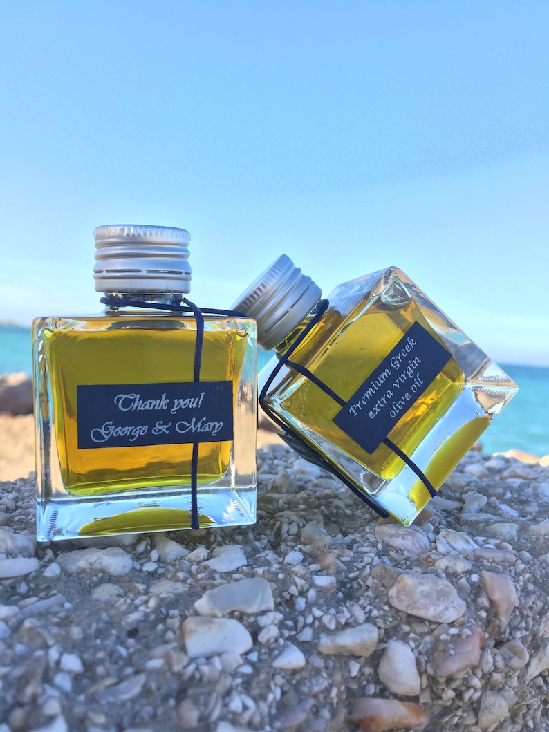Pode incluir: Duas pequenas garrafas de vidro de azeite de oliva com tampas prateadas. As garrafas est&atilde;o sentadas em uma superf&iacute;cie rochosa. Uma garrafa tem um r&oacute;tulo que diz "Obrigado! George & Mary". A outra garrafa tem um r&oacute;tulo que diz "Azeite de oliva extra virgem grego premium".