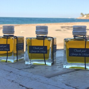 Pode incluir: Tr&ecirc;s pequenas garrafas de vidro cheias de azeite de oliva, cada uma com um r&oacute;tulo preto que diz "Obrigado! George & Mary". As garrafas est&atilde;o sobre uma superf&iacute;cie de madeira com uma praia de areia e o oceano ao fundo.