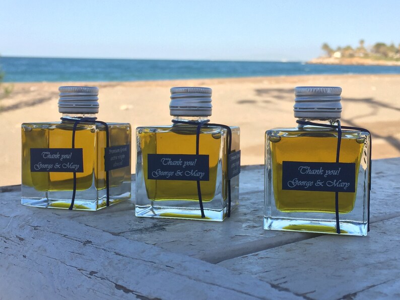 Pode incluir: Tr&ecirc;s pequenas garrafas de vidro cheias de azeite de oliva, cada uma com um r&oacute;tulo preto que diz "Obrigado! George & Mary". As garrafas est&atilde;o sobre uma superf&iacute;cie de madeira com uma praia de areia e o oceano ao fundo.