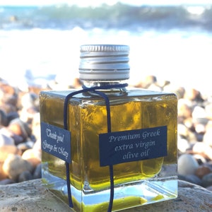 Pode incluir: Uma garrafa quadrada de vidro de azeite extra virgem grego premium com uma tampa prateada e uma fita preta. A garrafa est&aacute; sentada em uma rocha com um fundo desfocado de uma praia e o oceano.