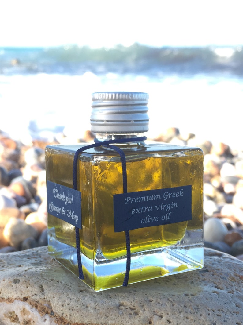 Pode incluir: Uma garrafa quadrada de vidro de azeite extra virgem grego premium com uma tampa prateada e uma fita preta. A garrafa est&aacute; sentada em uma rocha com um fundo desfocado de uma praia e o oceano.