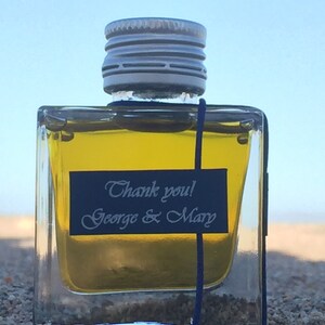 Pode incluir: Uma pequena garrafa de vidro transparente com uma tampa prateada e um r&oacute;tulo preto que diz "Obrigado! George & Mary". A garrafa est&aacute; cheia de um l&iacute;quido amarelo e est&aacute; sentada em uma superf&iacute;cie arenosa.