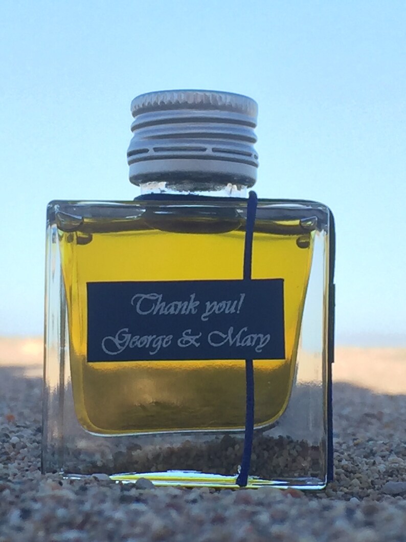 Pode incluir: Uma pequena garrafa de vidro transparente com uma tampa prateada e um r&oacute;tulo preto que diz "Obrigado! George & Mary". A garrafa est&aacute; cheia de um l&iacute;quido amarelo e est&aacute; sentada em uma superf&iacute;cie arenosa.