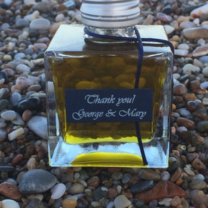 Pode incluir: Uma pequena garrafa de vidro transparente cheia de um l&iacute;quido dourado. A garrafa tem uma tampa prateada e uma fita preta amarrada ao redor do pesco&ccedil;o. Um r&oacute;tulo preto na garrafa diz "Obrigado! George & Mary".