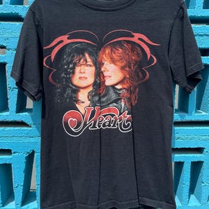 May include: Black t-shirt featuring a graphic of two women with long hair, framed by red decorative elements. The word "Heart" is written in a stylized font with a red heart. The shirt is hanging on a black hanger against a blue brick wall.