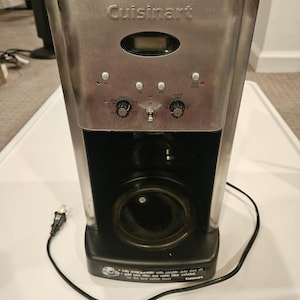 Peut inclure: Une cafetière Cuisinart en acier inoxydable et noire. Elle dispose d'un écran numérique, de boutons de commande et d'un cordon d'alimentation. Le texte sur la machine comprend "Cuisinart" et "fully programmable with variable auto shut off".