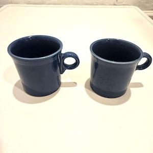 Puede incluir: Dos tazas de cerámica azul oscuro a juego con un acabado liso, cada una con un borde ancho ligeramente acampanado y una pequeña asa circular unida al lateral. Las tazas tienen una forma cilíndrica sencilla y un color sólido y uniforme.