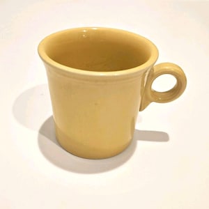Puede incluir: Una taza de cerámica de color amarillo claro sólido con un acabado suave y brillante. La taza presenta un borde ancho y acampanado y un asa circular pequeña unida al lateral. El diseño es sencillo y funcional, con un color amarillo pálido uniforme.