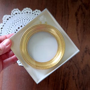 Könnte beinhalten: Ein kleiner, runder, goldfarbener Bilderrahmen mit weißem Innenraum, präsentiert in einer quadratischen, cremefarbenen Box. Der Rahmen hat ein detailliertes, strukturiertes Design. Die Box wird von einer Hand gehalten, mit einer weißen Spitzendecke im Hintergrund.