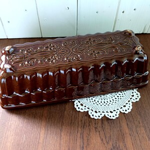 Könnte beinhalten: Eine braune Keramik-Kastenform mit einem dekorativen Blumendesign auf der Oberseite. Die Form hat eine geriffelte Außenseite und liegt auf einer weißen Spitzendecke. Die Form steht auf einer Holzoberfläche.