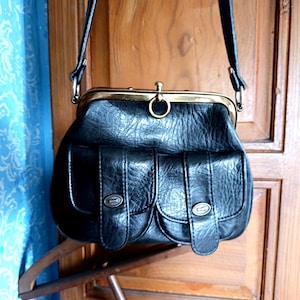 Puede incluir: Bolso vintage negro con cierre de broche y dos bolsillos delanteros. El bolso tiene un aspecto texturizado y una correa larga para el hombro. Los herrajes metálicos son dorados. El bolso está colgado de una percha de madera.