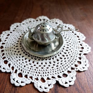 Può includere: Un piccolo infusore da tè color argento a forma di teiera è appoggiato su un piattino abbinato. L'infusore e il piattino poggiano su una tovaglietta bianca all'uncinetto con bordo smerlato. Il tutto è su una superficie di legno scuro.