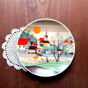 May include: A decorative plate with a village scene, including a church, houses, and figures in a field. The plate has a white rim and a colourful illustration with red, orange, green, and brown hues. The plate is placed on a white lace doily.