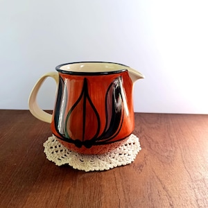 Puede incluir: Una jarra de cerámica con asa y boquilla crema, con un diseño floral negro y naranja. La jarra descansa sobre un tapete de ganchillo crema, sobre una superficie de madera.