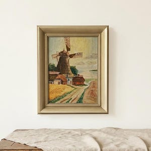 May include: A framed oil painting depicting a landscape with a central windmill, small houses with red roofs, a dirt path, and golden fields under a pale sky. The artwork is enclosed in a wide, metallic gold-toned frame with a beveled edge.