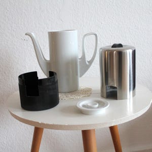 Può includere: Una caffettiera in ceramica bianca con beccuccio e manico lunghi, un contenitore in acciaio inossidabile, un supporto nero e un piattino bianco sono su un tavolo bianco con gambe in legno.