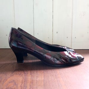 Puede incluir: Un par de zapatos de tacón bajo de color burdeos con un acabado brillante y texturizado. Los zapatos tienen una punta afilada y un tacón pequeño y oscuro. El fondo es una pared de madera clara y una superficie de madera.