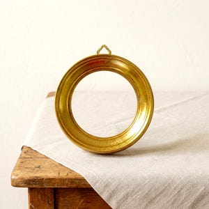 May include: A circular picture frame with a metallic gold finish and a smooth, rounded profile. The frame features a small, triangular hanging loop attached to the top edge, designed for wall mounting. The surface has a reflective sheen.