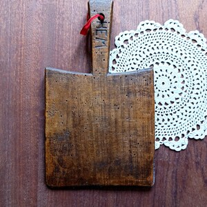 Puede incluir: Tabla de cortar rectangular de madera marrón oscuro con un asa grabada con la palabra "HEM". Una cinta roja está atada al asa. Un mantelito de encaje blanco se encuentra en el fondo.