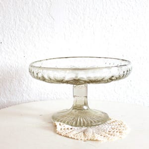 Puede incluir: Un soporte para pasteles de vidrio transparente con una parte superior redonda y una base decorativa. El soporte es de vidrio transparente y se encuentra sobre una superficie blanca. El diámetro del soporte es de aproximadamente 15 cm.