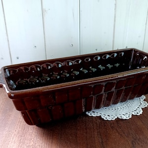 Puede incluir: Un molde para pan rectangular de cerámica marrón con acabado brillante. El molde presenta un diseño decorativo texturizado en los laterales y un borde festoneado. Tiene un asa en un extremo y está sobre un tapete de encaje blanco.