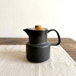 Könnte beinhalten: Eine mattschwarze Keramik-Teekanne mit zylindrischem Körper, einem geschwungenen Griff und einem kurzen Ausguss. Die Teekanne hat einen kontrastierenden, glänzend beigen Deckel mit einem kleinen Knauf in der Mitte. Das Design ist schlicht und glatt.