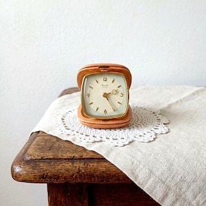 May include: A square analog alarm clock with a tan leather-like casing and a gold-toned frame. The clock face is off-white with gold-toned numerals at the 3, 6, 9, and 12 positions, gold-toned hands, and the brand name Kienzle printed in black.