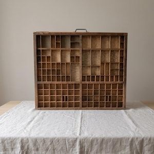 May include: A rectangular wooden printer's tray featuring a grid of numerous small, open compartments of varying sizes. The natural brown wood shows visible grain and texture, and a single metal handle is attached to the top center edge.