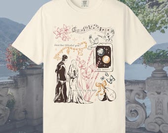 May include: Cream-colored t-shirt featuring a romantic illustration. The design includes a couple, flowers, butterflies, musical notes, and the text "love has blinded you?" and "meet me by the lake". The shirt has a classic crew neck and short sleeves.