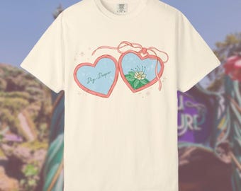 Op de afbeelding: Crèmekleurig T-shirt met een hartvormig ontwerp. Het ontwerp toont twee harten met een lichtblauwe achtergrond en roze randen. Het linkerhart heeft de tekst "Dig Deeper", en het rechterhart een bloemenillustratie. Een roze lint verbindt de harten.