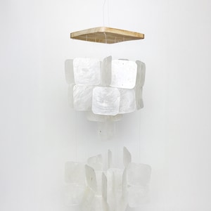 May include: A hanging light fixture with a square wooden top and multiple tiers of square, iridescent, shell-like pieces. The fixture is suspended by thin wires against a white background.