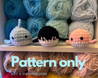 May include: Three crocheted whale toys in light blue, black and white, and pink and white, with small black eyes and fins. The toys are displayed on a shelf in front of a backdrop of various shades of blue yarn. Text on the shelf reads "Pattern only, NOT A FINISHED ITEM."
