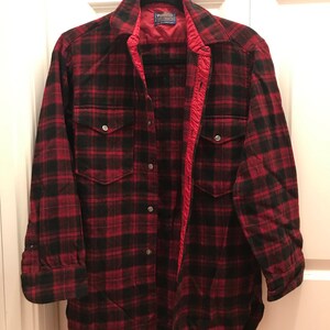 May include: A red and black plaid flannel shirt with a button-up front and long sleeves. The shirt has a label that reads "Penfield" and "Wool"