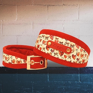 Peut inclure: Deux colliers pour animaux de compagnie rouges et crème. Les colliers présentent un extérieur en suédine rouge et un intérieur crème avec un motif de cœurs et de nœuds rouges. Un collier a une boucle dorée. Le nom "Ziggy" est imprimé sur une étiquette en cuir rouge.