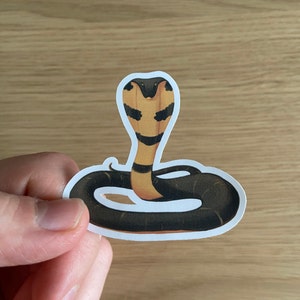 May include: A sticker of a black and yellow cobra with its hood raised, the snake is curled in a circle.